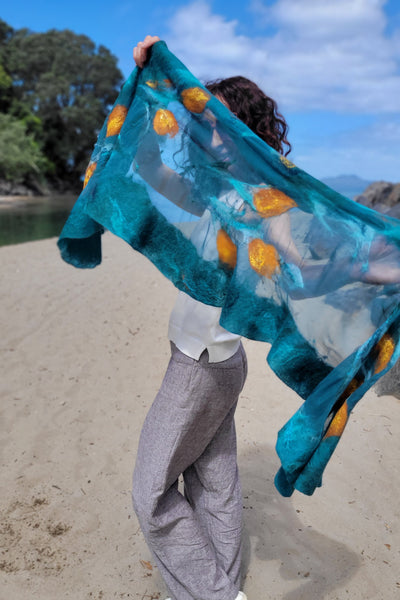 A woman wearing a handmade teal merino wool and silk shawl with golden kōwhai flowers on a New Zealand beach.