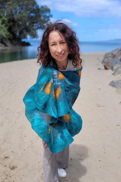 A woman with curly brown hair stands on a sandy beach wearing a vibrant teal and orange patterned wrap. Trees, rocks, and the ocean are visible in the background under a blue sky.