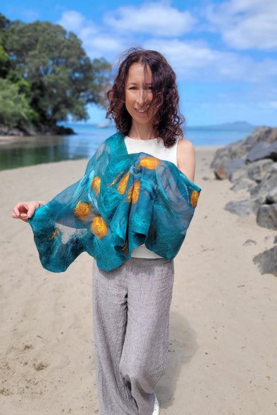 A woman standing on a sandy beach wearing a teal blue and yellow patterned shawl draped over her shoulders.