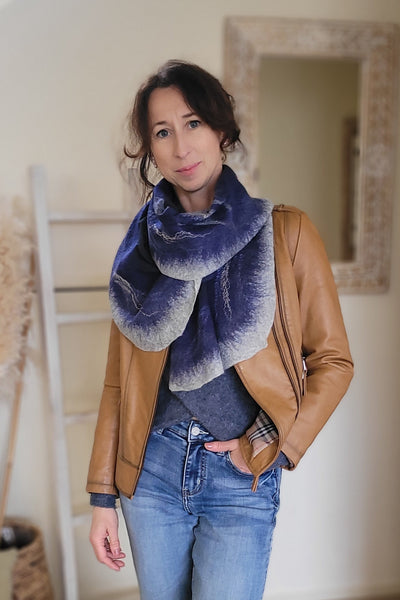A woman in a stylish outfit, including a crafted merino and silk scarf, a brown leather jacket, and blue jeans, is standing indoors. 