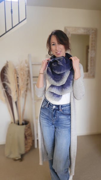 A woman standing in a room with a neutral colour scheme, wearing a wool scarf and long coat and jeans.