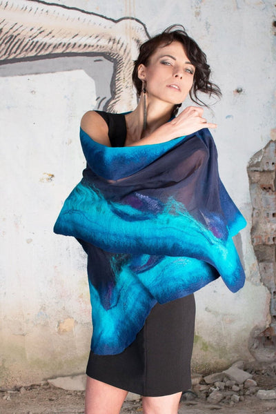 A woman wearing a blue and purple patterned scarf stands in front of a weathered concrete wall, with a sketch-like drawing on the wall behind her.