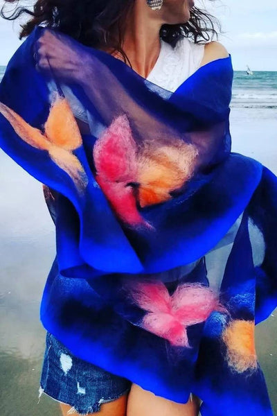 A woman wearing a blue and pink floral shawl stands on a beach, with the ocean visible in the background.