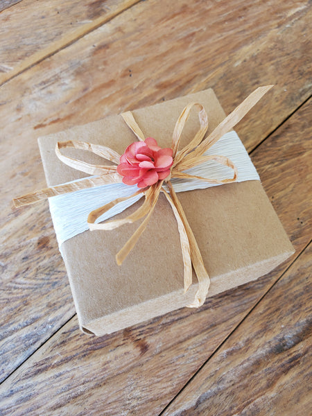 A brown gift box with a pink flower tied with a white ribbon, placed on a wooden surface.