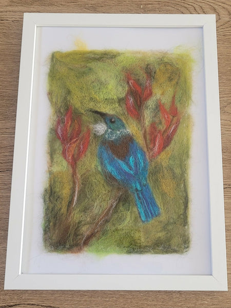 Felted artwork of a blue bird on a green background with red flowers, framed in white.