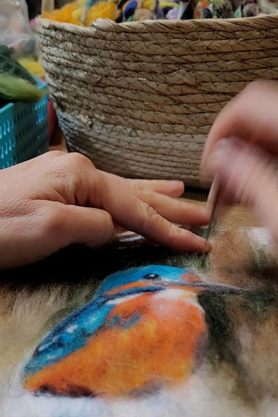 Person painting with wool a colourful bird on fabric with a woven basket in the background