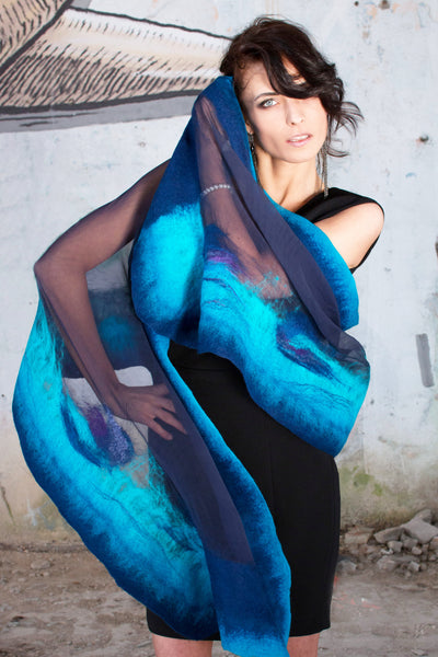 A woman wearing a black dress and a blue and purple scarf stands in front of a concrete wall, with her hair blowing in the wind.