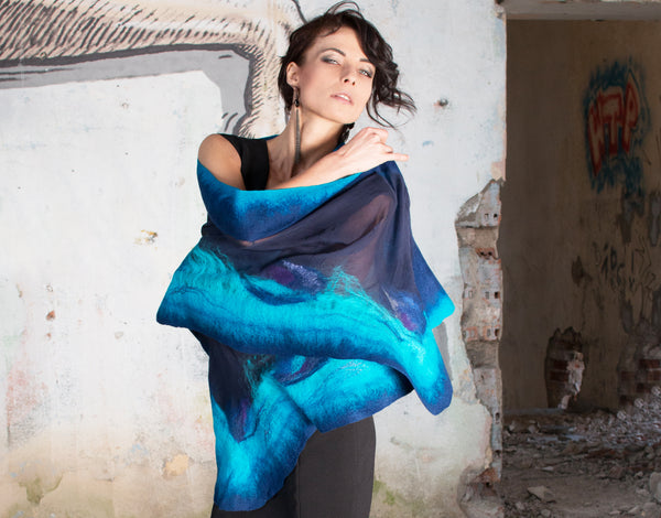 A woman stands in front of a dilapidated wall, wearing a blue and purple scarf.