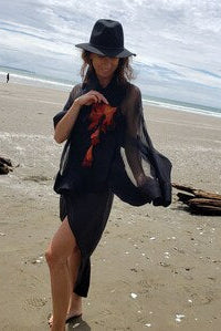 A woman stands on a sandy beach wearing a black wide-brimmed hat, a sheer black wrap, and a black shawl, with ocean and cloudy sky in the background.
