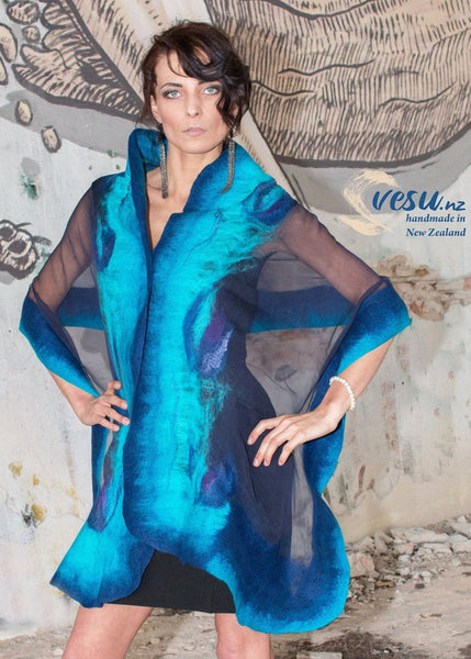 A woman wearing a blue and black patterned dress stands in front of a wall with a mural.