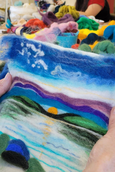 Colourful felted landscape with hands holding it, blurred background of more yarn.