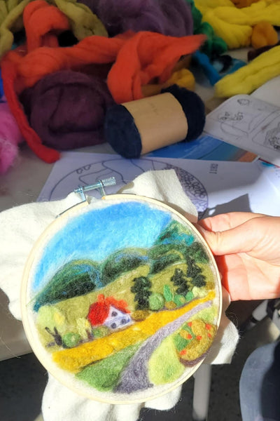 Needle felting landscape design, held by a hand, surrounded by colourful yarn and fabric.