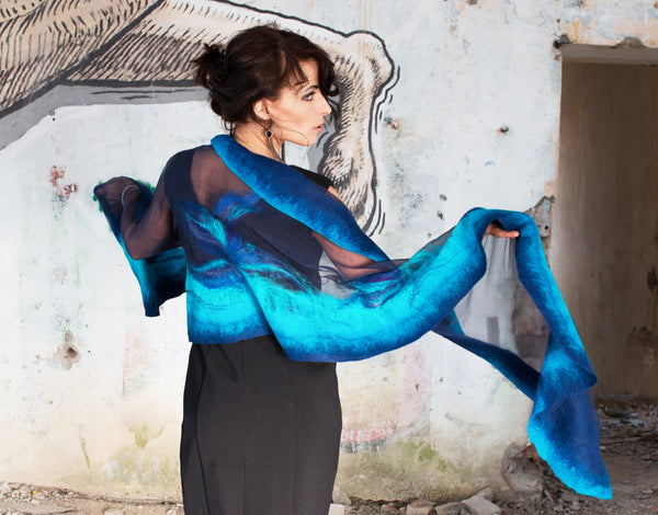 A woman in a black dress stands in front of a graffiti-covered wall, holding a blue scarf.