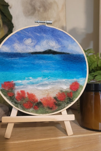Wool Painting in anEmbroidery hoop with a scenic design of a beach and red flowers on a wooden stand.