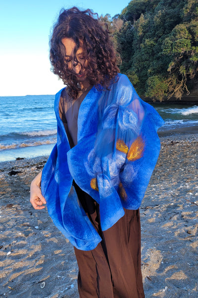 Handmade royal blue merino wool and silk floral shawl with golden accents, worn on a beach in New Zealand. VESU wearable art.