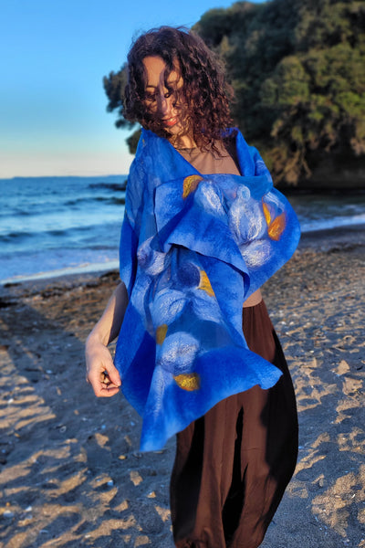 A woman stands on a sandy beach with the ocean and a rocky cliff in the background, wearing royal blue shawl with white and gold painted floral motifs over dark brown harem pants and white sneakers.